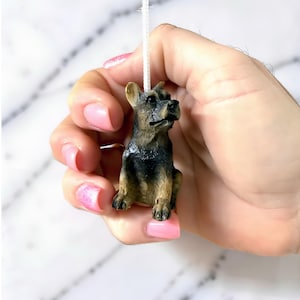 Könnte beinhalten: Eine kleine, detaillierte Figur eines Deutschen Schäferhundes. Der Hund hat ein schwarz-braunes Fell und sitzt. Er ist an einer weißen Schnur befestigt, was darauf hindeutet, dass es sich um einen Anhänger handelt.