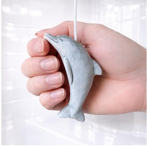 Op de afbeelding: Een lichtgrijze dolfijnvormige trekschakelaar voor een lamp. De dolfijn wordt door een hand tegen een witte achtergrond gehouden. De trekketting is aan de rug van de dolfijn bevestigd.