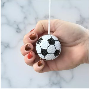 May include: A miniature soccer ball ornament, with a classic black and white design, suspended by a white string. The ornament is held in a hand with painted nails. The background is a white marble effect.