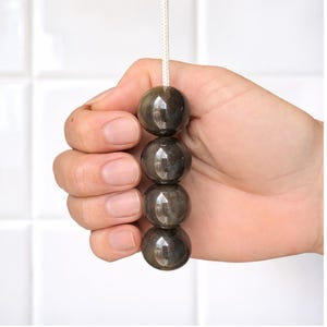 Puede incluir: Un conjunto de cuatro cuentas esféricas de color gris oscuro ensartadas en un cordón color crema. Las cuentas se sostienen en una mano, sobre un fondo de azulejos blancos.