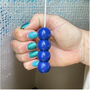 Könnte beinhalten: Eine Hand hält eine blaue Perlenzugkette. Die Perlen sind rund und glatt. Die Hand trägt blauen und grünen Nagellack.