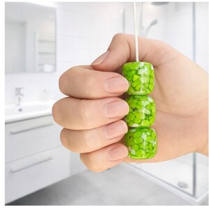 May include: A decorative light pull with three stacked, square green beads. The beads have a speckled pattern and are strung on a white cord. The pull is held in a hand, with a bathroom setting in the background.
