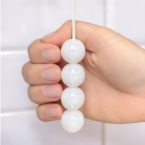 以下が含まれることがあります： 白く丸いビーズが4つ連なったプルチェーン。照明器具や扇風機などに使用される。白い紐で繋がれており、手で持たれている。背景は白で、シンプルなデザイン。