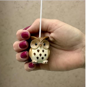 May include: A small, decorative owl-shaped light pull. The owl is cream-colored with brown accents and black spots. It is held by a hand with red nail polish, and a white cord extends from the top.