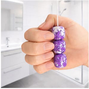 May include: A decorative bathroom light pull featuring three cube-shaped beads. The beads are a mix of purple and white, strung together on a white cord. The pull is held in a hand, with a bathroom in the background.