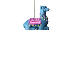 May include: A blue llama ornament with a pink and multi-colored saddle. The llama is sitting down with its head turned to the right.