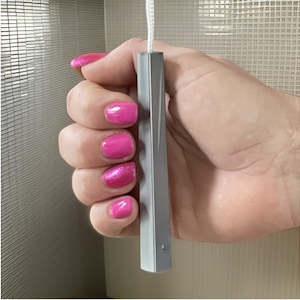 May include: A silver metal pull cord for window blinds. The cord is held in a hand with pink nail polish.