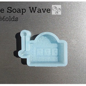 May include: A light blue silicone mold shaped like a slot machine with three windows. The mold is designed for making soap.