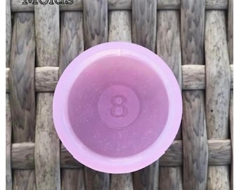 May include: A light purple silicone mold shaped like a pool ball with the number 8 printed on it. The mold is sitting on a woven surface.