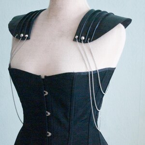 May include: Black corset with attached shoulder armor. The corset features a structured bodice with a series of metal clasps down the front. Silver chains drape from the shoulder armor to the corset. The shoulder armor is made of layered black material.