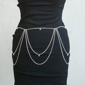 May include: A black sleeveless dress with a textured fabric and a horizontal cutout at the chest. A silver-toned metal body chain with multiple draped layers and small circular accents is worn around the waist, hanging down over the hips.