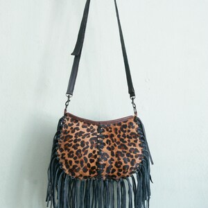 May include: A crossbody bag with a leopard print design. The bag features a black leather strap and fringe along the bottom. The bag is hanging against a white wall.