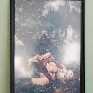 May include: Framed gothic art print featuring a woman in a burgundy dress lying in a graveyard. The artwork includes a castle, tombstones, and the artist's signature, "Victoria Frances."