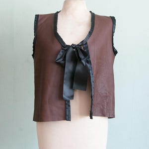 以下が含まれることがあります： 黒いトリムと黒いサテンのリボンが付いた茶色のベスト。ノースリーブのベストは、革または革のような素材で作られているようです。マネキンに飾られたベストです。