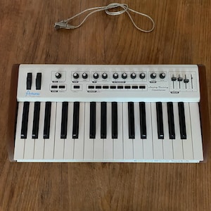 May include: An Arturia Analog Factory Experience MIDI controller with a white keyboard, black keys, and wood-toned side panels. The top panel features various knobs, sliders, and buttons, with a USB cable positioned above the device.