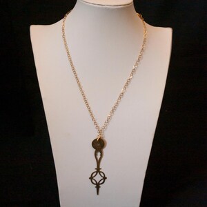Può includere: Una collana color oro con un ciondolo a forma di chiave. La collana è esposta su un manichino bianco su sfondo nero. Il ciondolo a chiave ha una parte superiore circolare e un design traforato decorativo.