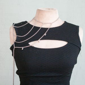 May include: A silver-toned metal body chain draped over the right shoulder and across the chest, featuring multiple delicate linked chains and small dangling charms, worn over a black sleeveless top with a horizontal cutout across the chest.