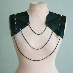 May include: A black shoulder piece with silver spikes and chain detailing. The accessory is displayed on a mannequin, showcasing its geometric design and edgy aesthetic. The piece features multiple rows of delicate chains.