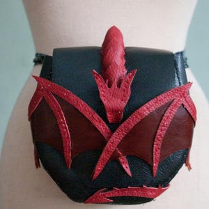 Op de afbeelding: Een zwarte leren tas met een rode drakenapplicatie op de voorflap. Het drakenontwerp bevat gestructureerde rode leren vleugels, een centrale kop en een staartdetail aan de onderkant. De tas wordt vastgemaakt met een riem om de taille.