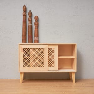 Puede incluir: Un mueble de madera clara con una puerta corredera con diseño de celosía. Tres figuras de madera tallada se alzan encima. El mueble tiene un estante y está sobre patas anguladas. El fondo es una pared gris claro.