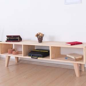 May include: A light-colored wooden TV stand with four cubby spaces and angled legs. The stand holds a red abacus, a black case, a red book, and two small candles. A potted plant is to the left.