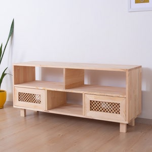 May include: A light-coloured wooden TV stand with two doors featuring a lattice design, open shelving, and a rectangular shape. The stand is supported by short, square legs. A tall green plant in a yellow pot is to the left.