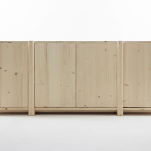 May include: A light-coloured wooden cabinet with four doors and a rectangular shape. The cabinet is made of unfinished wood, showcasing the natural grain and knots. The design features vertical supports on each side and between the doors.