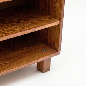 May include: A wooden bookshelf with two visible shelves. The shelves and sides are made of a warm-toned wood, showcasing a prominent wood grain pattern. The bookshelf has a simple, sturdy design, with a rectangular base.