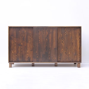 May include: A dark brown wooden cabinet with sliding doors. The cabinet has a rectangular shape and is supported by short, square legs. The wood grain is visible, adding texture to the piece. The cabinet is against a white background.