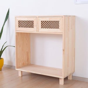 Puede incluir: Un mueble de madera clara con dos puertas superiores con diseño de celosía. El mueble tiene un estante abierto debajo y se apoya en cuatro patas cortas. La veta de la madera es visible y el mueble está contra una pared blanca.