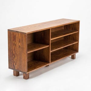 May include: A wooden bookshelf with a warm brown finish. The bookshelf has multiple shelves for storage and is supported by short, square legs. The wood grain is visible, adding to its natural aesthetic. The design is simple and functional.