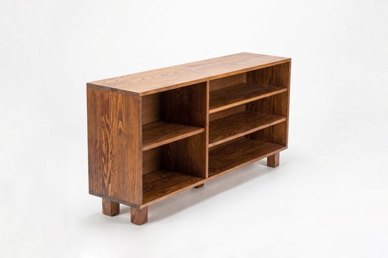 May include: A wooden bookshelf with a warm brown finish. The bookshelf has multiple shelves for storage and is supported by short, square legs. The wood grain is visible, adding to its natural aesthetic. The design is simple and functional.