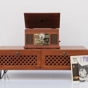 May include: A vintage wooden radio and record player combo unit with a dark brown finish. The unit sits atop a matching cabinet with latticework doors. A vinyl record is propped up next to the cabinet. The radio has a brass-colored face.