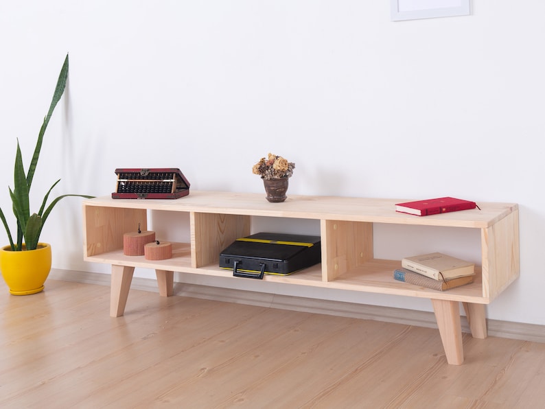 May include: A light-colored wooden TV stand with four cubby spaces and angled legs. The stand holds a red abacus, a black case, a red book, and two small candles. A potted plant is to the left.