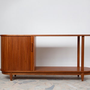 May include: A wooden cabinet in a mid-century modern style, finished in a warm brown tone. It has a slatted door on the left, a long tabletop, and a lower shelf. The design incorporates rounded edges and tapered legs, blending functionality with style.