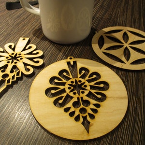 May include: A collection of three laser-cut wooden ornaments with intricate designs. One is a teardrop shape, another is circular, and the third is a unique floral design. A white mug is in the background. The ornaments are made of light-colored wood.