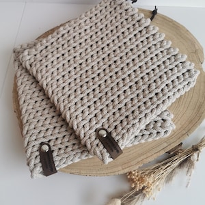 Könnte beinhalten: Zwei beige, handgehäkelte Topflappen mit einem strukturierten Webmuster. Jeder Topflappen hat ein kleines braunes Lederetikett. Die Topflappen werden auf einer Holzscheibe mit getrockneten Blumen im Hintergrund präsentiert.