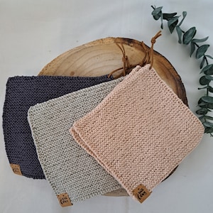 Könnte beinhalten: Drei gestrickte Waschlappen in Grau-, Beige- und Rosatönen sind auf einer Holzoberfläche angeordnet. Jeder Lappen hat ein kleines braunes Etikett mit dem Aufdruck "with love". Ein Lederriemen ist an der Holzoberfläche befestigt.