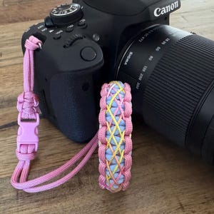 May include: A black Canon DSLR camera with a Sigma lens attached, featuring a pink paracord wrist strap with a plastic buckle and a matching woven paracord bracelet in pink, light blue, and yellow colours positioned in front of the camera body.