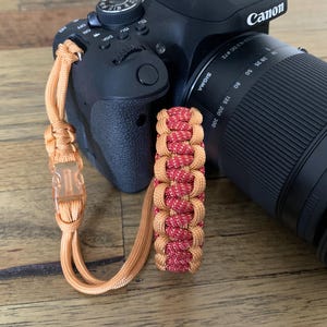 May include: A black Canon DSLR camera with a Sigma lens attached, featuring a braided paracord wrist strap. The strap is constructed from solid orange cord and a central section of interwoven red and orange cord, secured by a translucent orange plastic buckle.