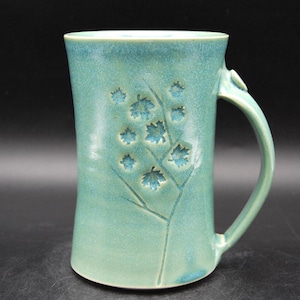 May include: A tall, turquoise ceramic mug with a large handle. The mug features an embossed design of leaves and branches. The mug is approximately 6 inches tall and has a smooth, glossy finish. The mug is suitable for hot or cold beverages.