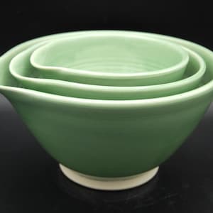 May include: A set of three nested ceramic mixing bowls in a soft sage green glaze. Each bowl features a smooth, rounded interior and a distinct pouring spout on one side. The bowls have a white circular base and a glossy, uniform finish.
