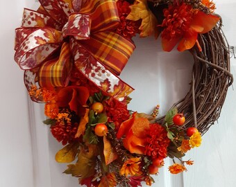 May include: A fall-themed wreath with a large orange and red plaid bow. The wreath is made of grapevine and is decorated with orange, red, and yellow flowers, leaves, and berries.