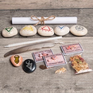 May include: A set of five white stones with painted symbols, a rolled up scroll, a white feather, a white candle, and four small bags with the text "Sea Witch Wisdom" and various items like shells, flowers, and a coin.