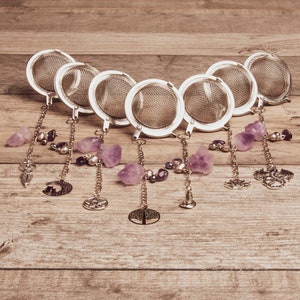 May include: Set of eight silver tea infusers with amethyst crystal charms and decorative metal pendants. The charms include a crescent moon, a tree, a star, a flower, a dragon, a heart, a crescent moon, and a star.