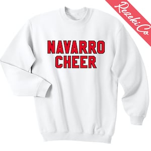 Puede incluir: Sudadera blanca de cuello redondo con las palabras "NAVARRO CHEER" en letras rojas en negrita con contorno negro. La sudadera tiene mangas largas y cuello y puños acanalados. La marca "Rezeki Co" está impresa en una franja diagonal roja.