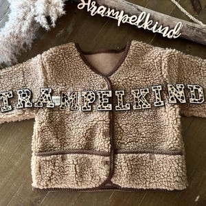 Könnte beinhalten: Eine beige, flauschige Babyjacke mit braunem Besatz und Knopfverschluss. Das Wort "STRAMPELKIND" ist in großen, dekorativen Buchstaben auf der Vorderseite geschrieben. Die Jacke hat ein weiches, strukturiertes Aussehen.