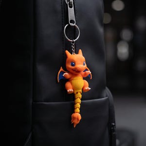 May include: An orange and yellow articulated dragon keychain with blue wing accents attached to a black backpack zipper. The text SkeletonEarth Designs, LEVEL UP YOUR EVERYDAY CARRY. The ultimate articulated fidget companion. Ready to attach and impossible to put down. is visible.