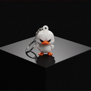 May include: A white duck keychain with an angry facial expression, featuring a bright orange beak and orange feet. The figure is attached to a silver-colored metal split ring and chain. The text Cure plain keychain fatigue with instant humor and personality. The ultimate conversation starter starter for your backpack or desk. is visible.