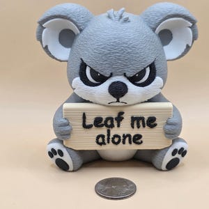 May include: A gray koala figurine with a textured surface, large ears, and an angry facial expression. The koala holds a rectangular sign with the text "Leaf me alone" in black lettering. A US nickel coin is placed in front for scale.
